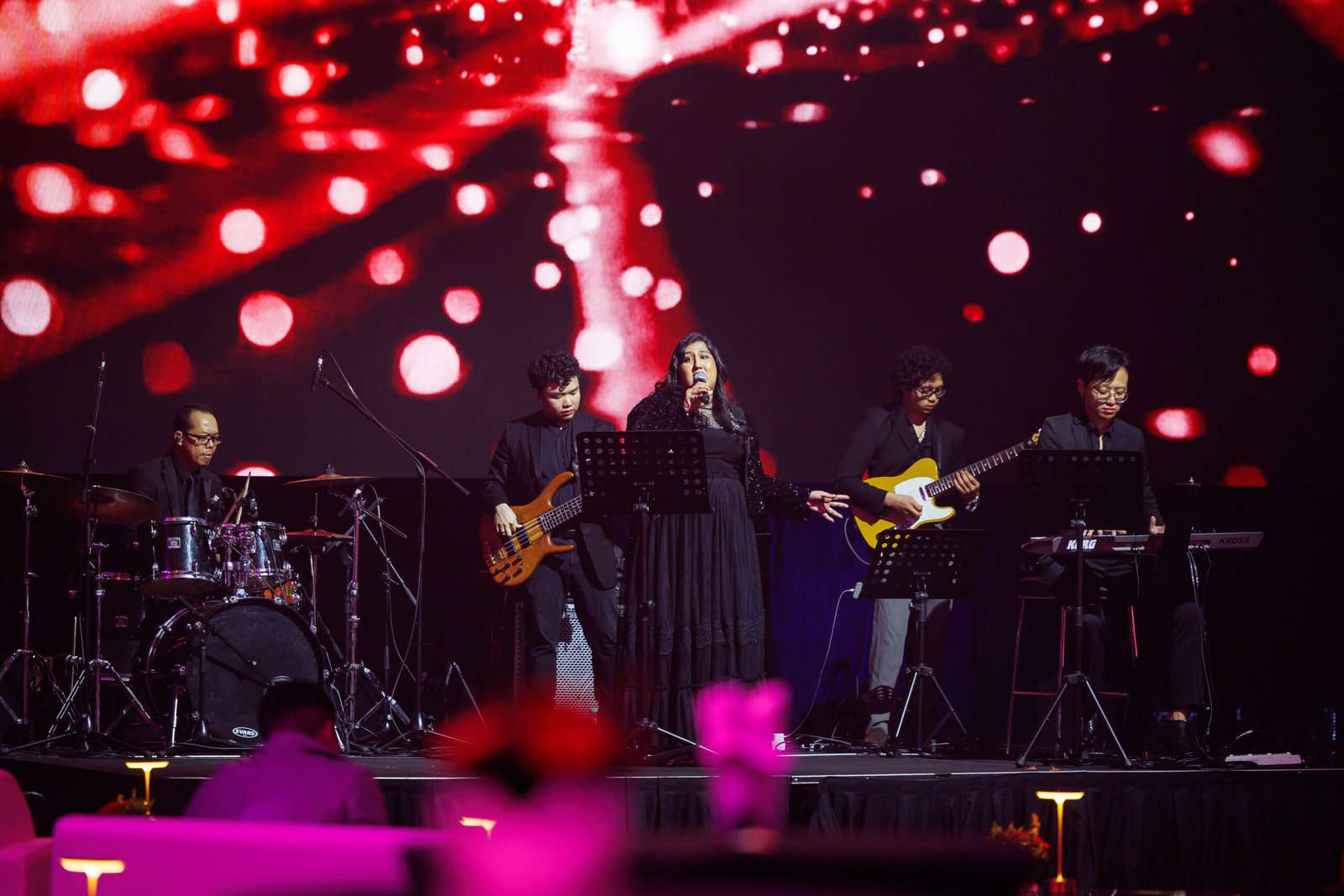 Live band performance on stage with dramatic red bokeh backdrop and pink table lighting; five musicians performing at gala